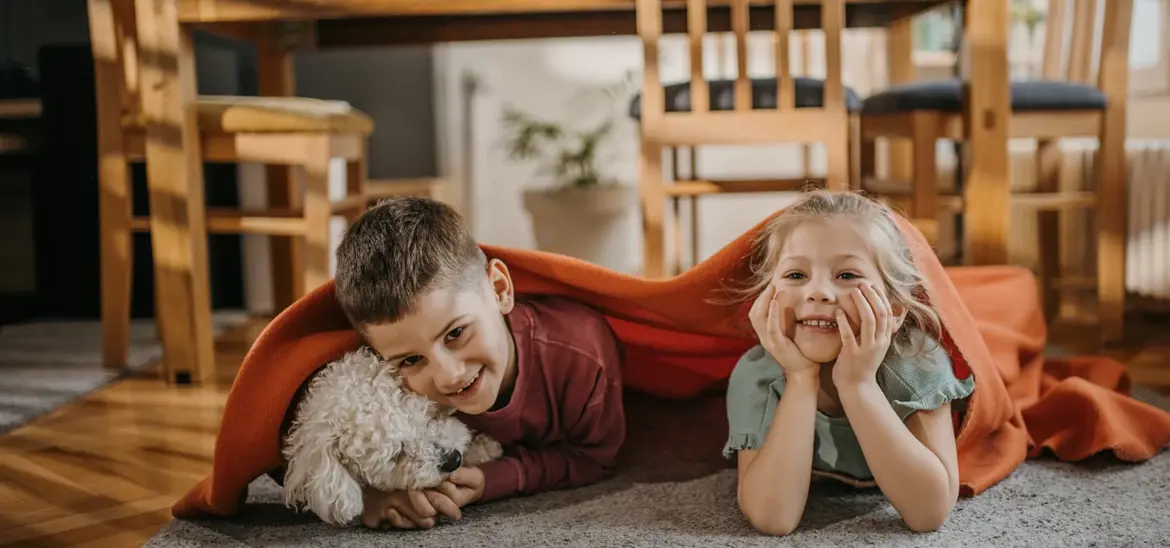 A boy and a girl lying on the floor, cuddling with a dog under a blanket