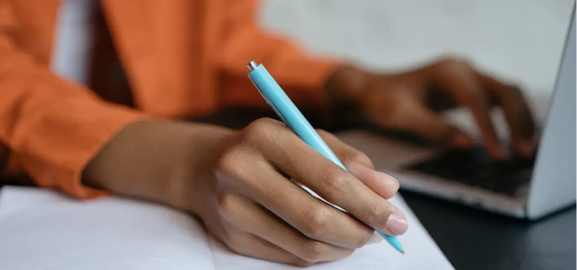 A woman writing on a notebook while looking at a laptop