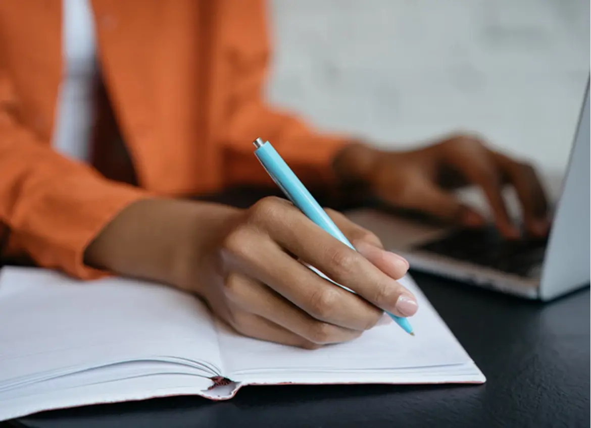 A woman writing on a notebook while looking at a laptop