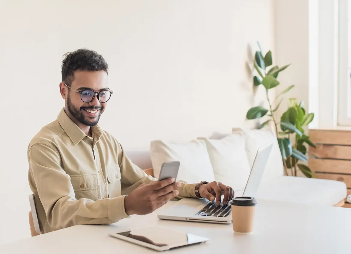 A man smiling viewing his phone while working on his laptop